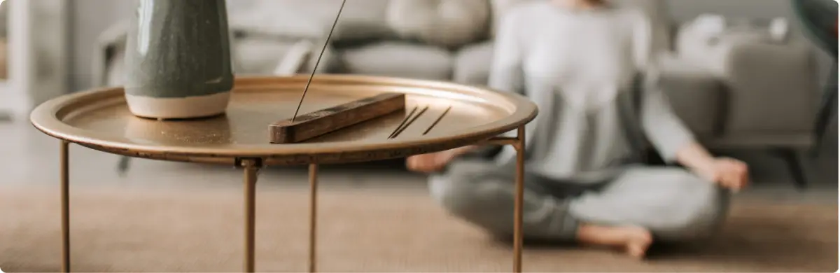 Burning incense on a table with a person meditating in the background for holistic support.